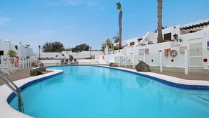 Une piscine avec une plage blanche, des palmiers et un bâtiment blanc surmonté d’un toit recouvert de tuiles bleues.