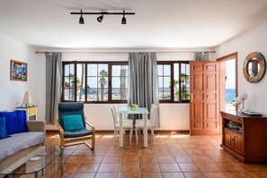 A room with a sofa, a dining table with chairs, a wooden cabinet, and a view of the sea through large windows.