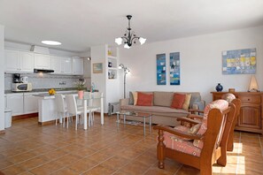 Eine moderne Küche mit weißen Schränken, ein Essbereich mit einem Glastisch, ein Sofa mit gemusterten Kissen und ein Holzstuhl.