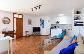 Una sala de estar con televisor, una zona de comedor con mesa y sillas, una cocina con microondas y cafetera, y un balcón pequeño.