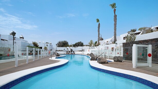 Une piscine à bordures courbes, entourée de bâtiments blancs agrémentés d’éléments en pierre, de palmiers et surplombée d’un ciel bleu azur.