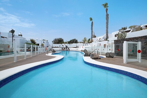 Une piscine à bordures courbes, entourée de bâtiments blancs agrémentés d’éléments en pierre, de palmiers et surplombée d’un ciel bleu azur.