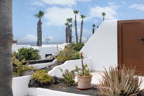 A white building with a brown door, surrounded by various plants and a view of the sea.