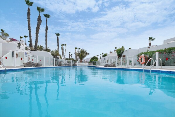 A clear blue swimming pool with white buildings and palm trees in the background.