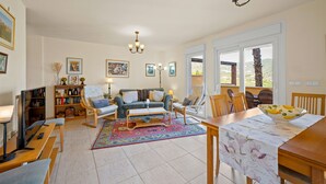 A living room with a sofa, coffee table, and dining table. There are bookshelves, a TV, and a chandelier. - Panoramic Sea View Villa with Large Terrace, Shared Pools & Space for 8 (Almunecar)