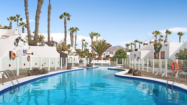 A swimming pool with clear blue water, surrounded by white buildings and palm trees. - Apartment "Puerto Chico 7" with Sea View, Shared Pool, Terrace & Wi-Fi (Playa Blanca)