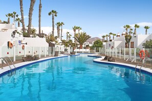 A swimming pool with clear blue water, surrounded by white buildings and palm trees.