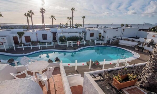 Une piscine entourée de bâtiments blancs, de palmiers, sous un ciel dégagé, avec une vue sur la mer.