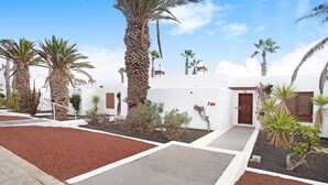 A courtyard with palm trees, a white building with a brown door, and a red and gray paved area. - Apartment "Puerto Chico 7" with Sea View, Shared Pool, Terrace & Wi-Fi (Playa Blanca)