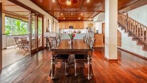 A spacious dining area with a large wooden table and chairs, a ceiling fan, and a view of the kitchen and outdoor area. - Camplung Beach Villa and Farmstead (Tejakula)