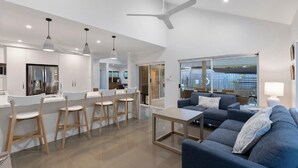 A modern kitchen with a refrigerator, bar stools, and pendant lights. There is a living area with a sofa and a coffee table.