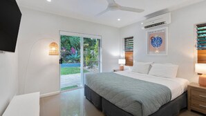 A modern bedroom with a large bed, a ceiling fan, a TV, and a view of a garden through a sliding glass door.
