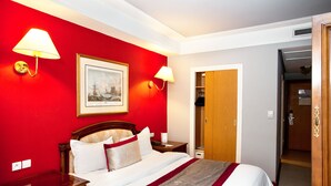 Una habitación de hotel con una pared de acento roja, una cama con ropa blanca, dos lámparas de noche, una mesita de noche de madera, un cuadro enmarcado y una puerta de madera que da a un armario.