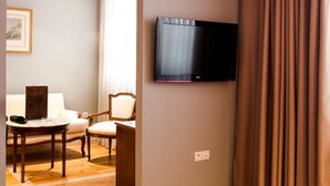 Habitación de hotel con cama, televisor montado en la pared, zona de comedor con mesa y sillas, y vista al pasillo.