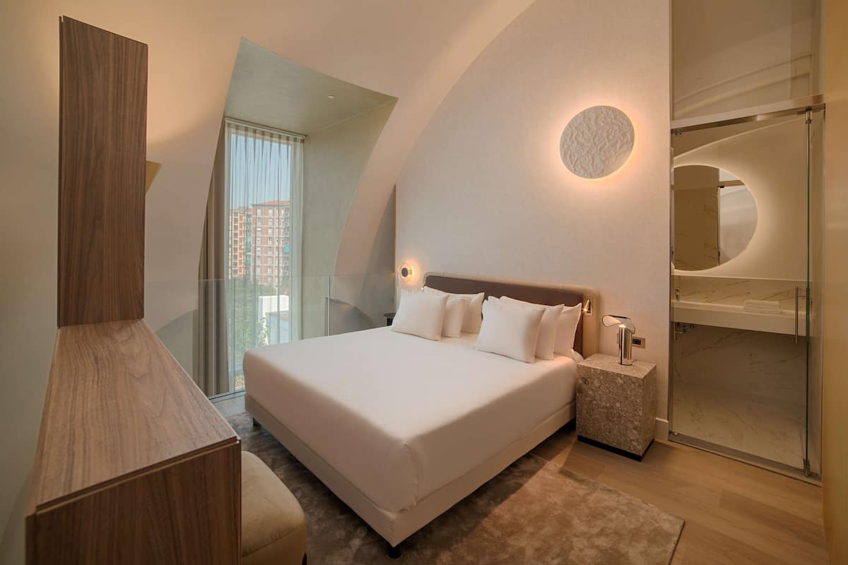 A modern bedroom with a large bed, a wooden desk, a glass shower enclosure, and a city view.