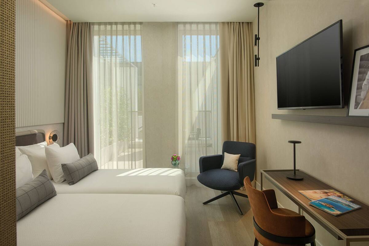 A hotel room with two beds, a TV, a desk with a chair, and large windows with curtains.