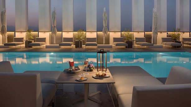 Ein Poolbereich mit Liegestühlen, einem Tisch mit Speisen und Getränken sowie hohen, vertikalen Lampen.