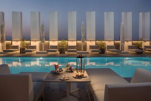 Uma área à beira da piscina com espreguiçadeiras, uma mesa com comida e bebidas, e painéis de luz verticais.