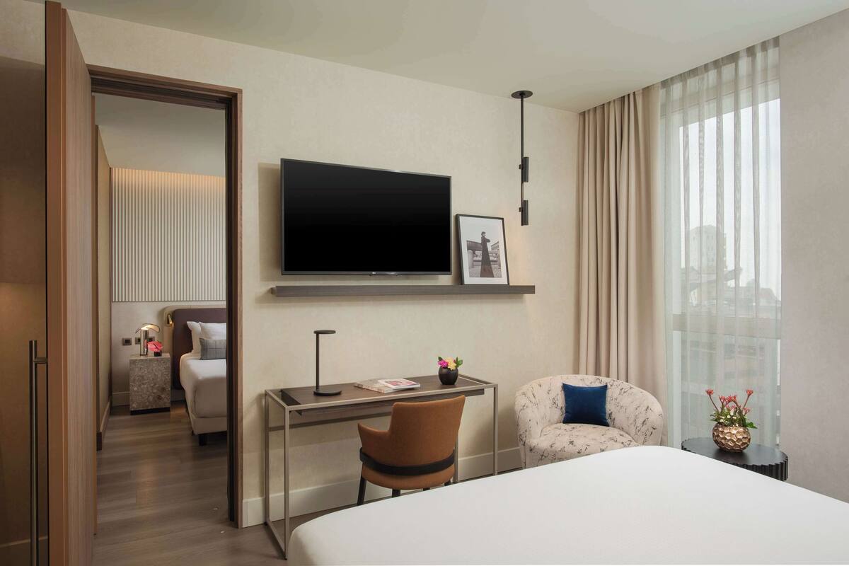 A hotel room with a bed, a desk with a chair, a TV mounted on the wall, and a window with curtains.