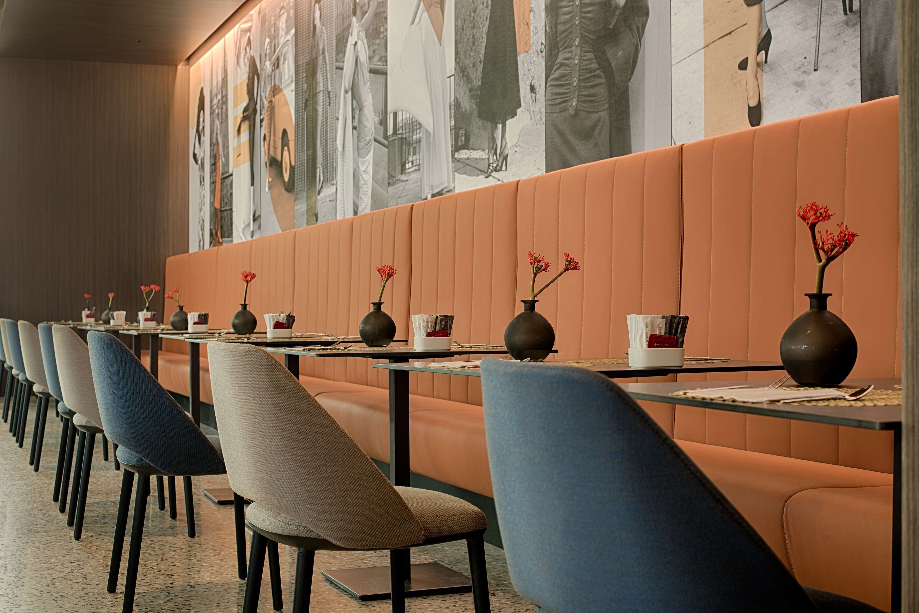 Uma fileira de mesas de jantar com cadeiras, cada mesa tem um vaso com flores vermelhas, e as paredes são decoradas com fotografias em preto e branco de grande formato.
