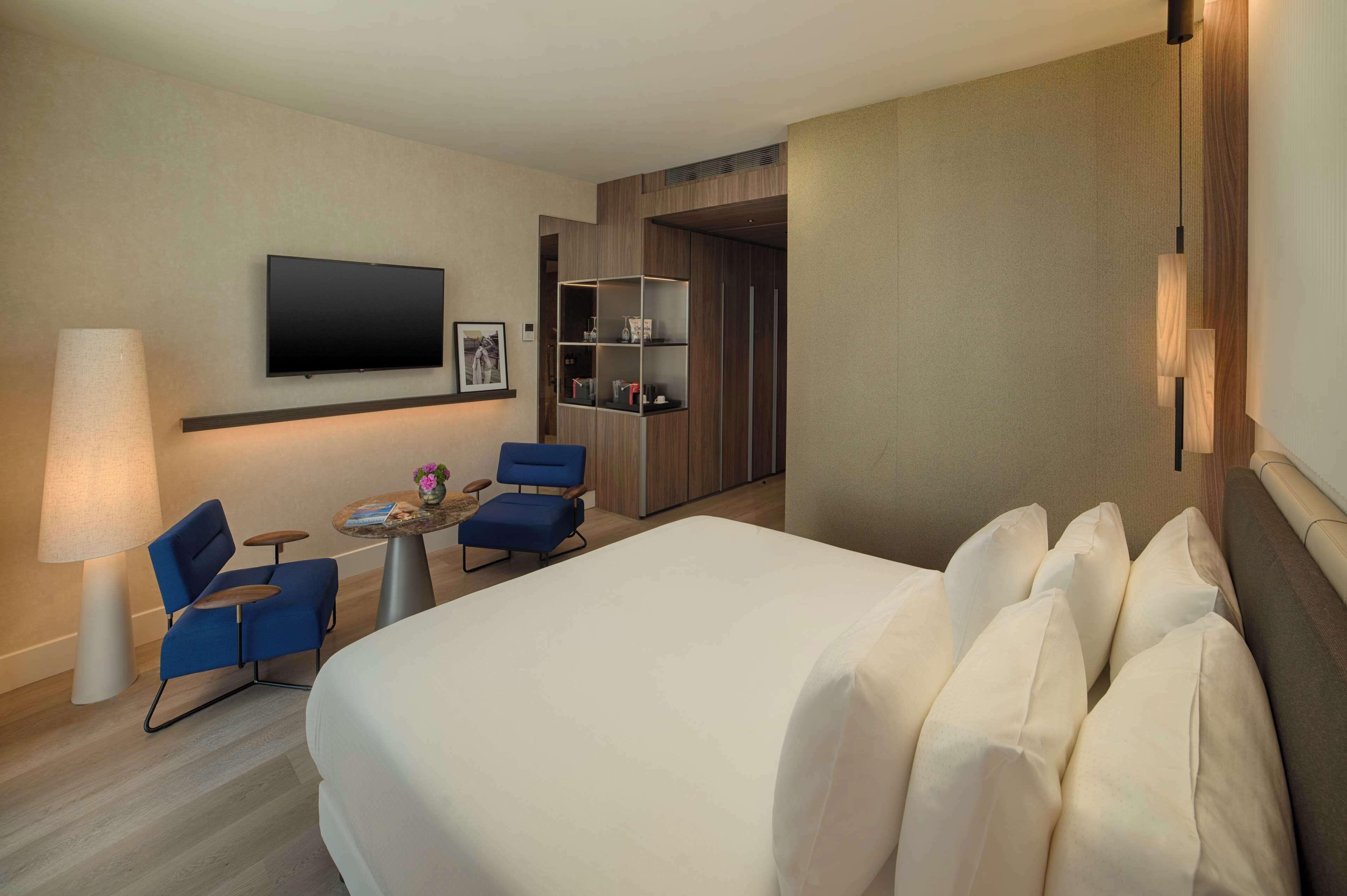 Ein Hotelzimmer mit einem großen Bett, einem an der Wand befestigten Flachbildfernseher, einem Schreibtisch mit Stuhl, einer Lampe und einem Bücherregal.
