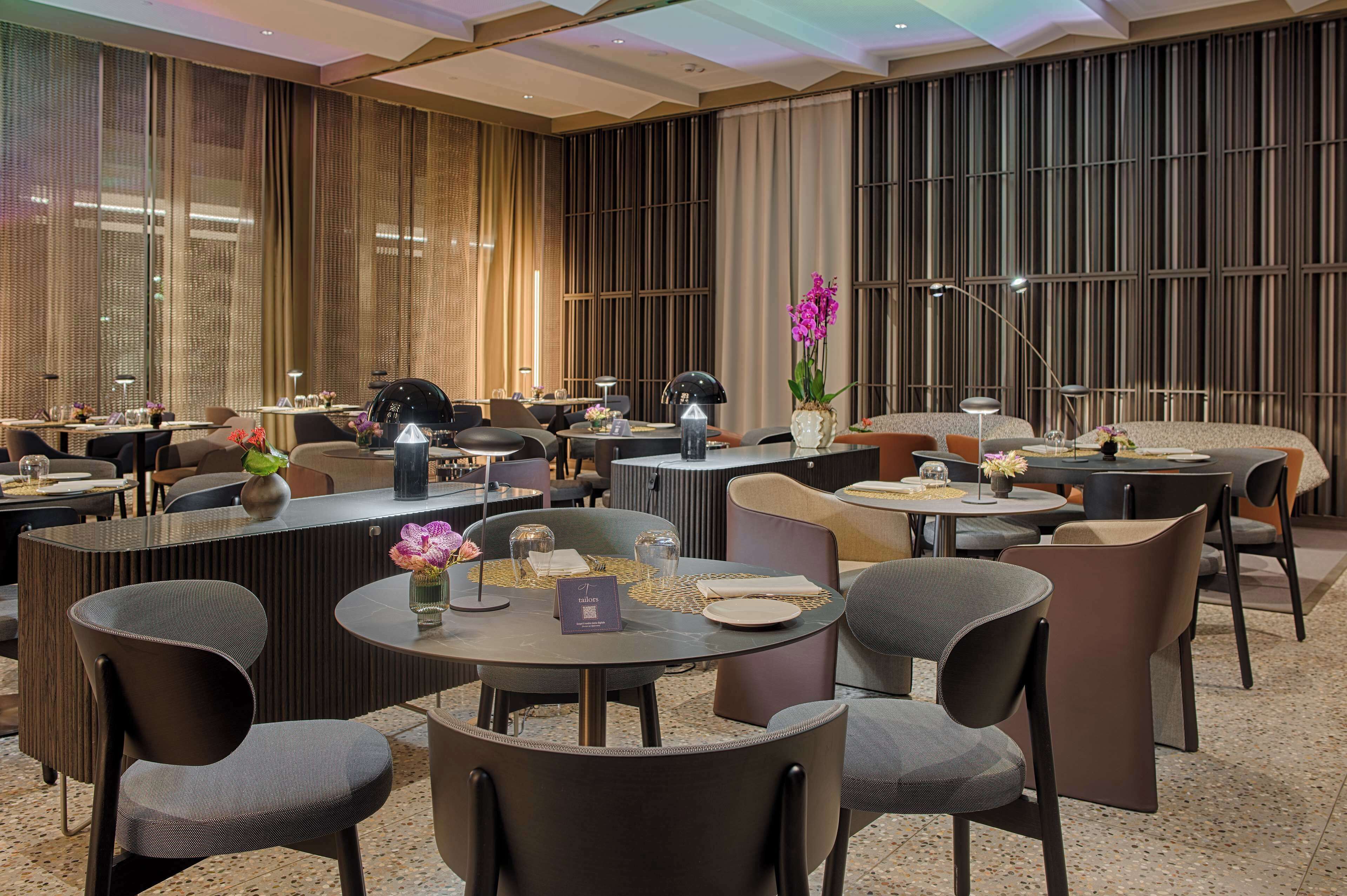 A modern hotel lobby with multiple seating areas, including round tables and chairs, and a reception desk.