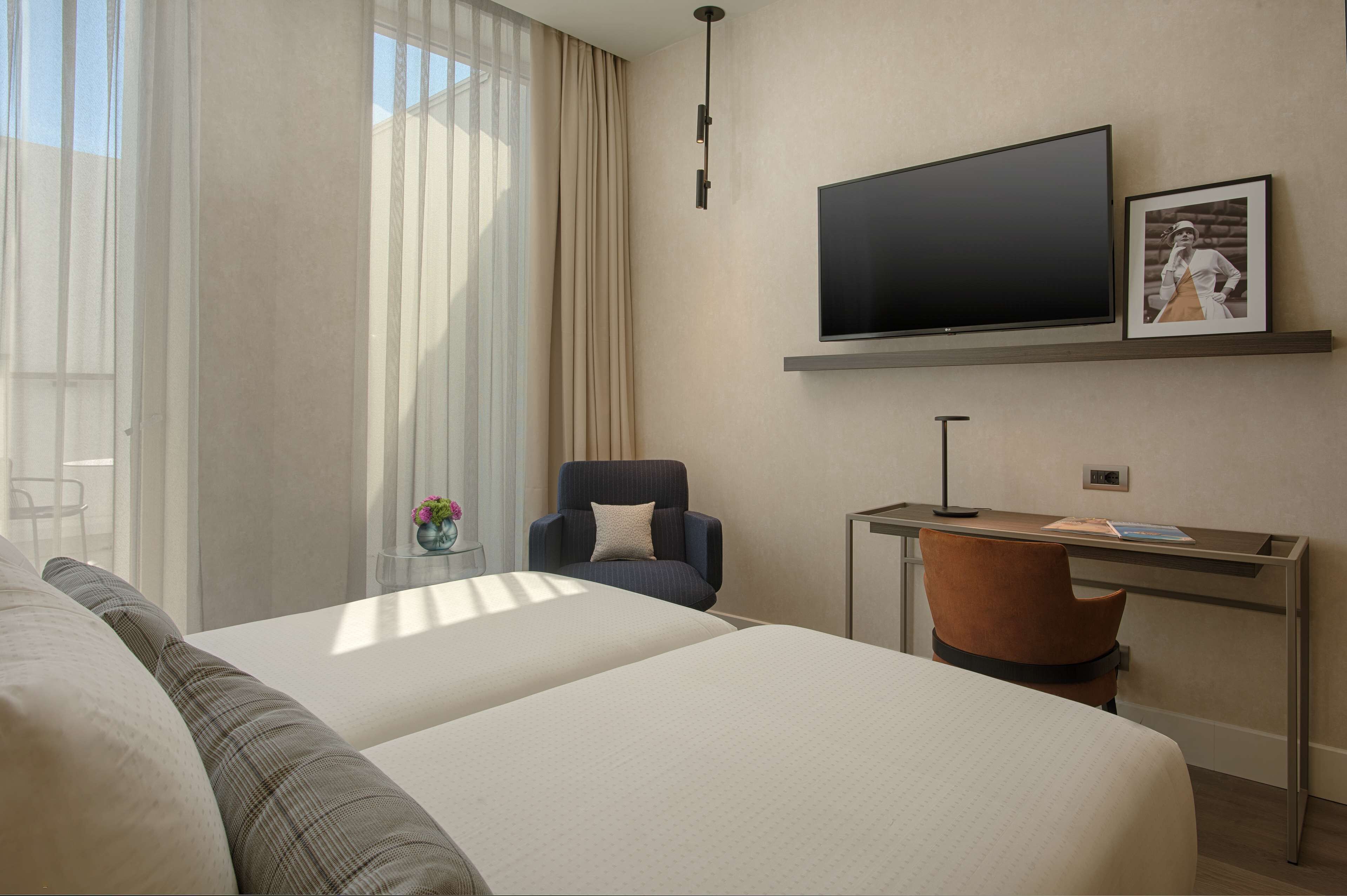 Ett hotellrum med en säng, ett skrivbord, en stol, en tv och ett fönster med gardiner.