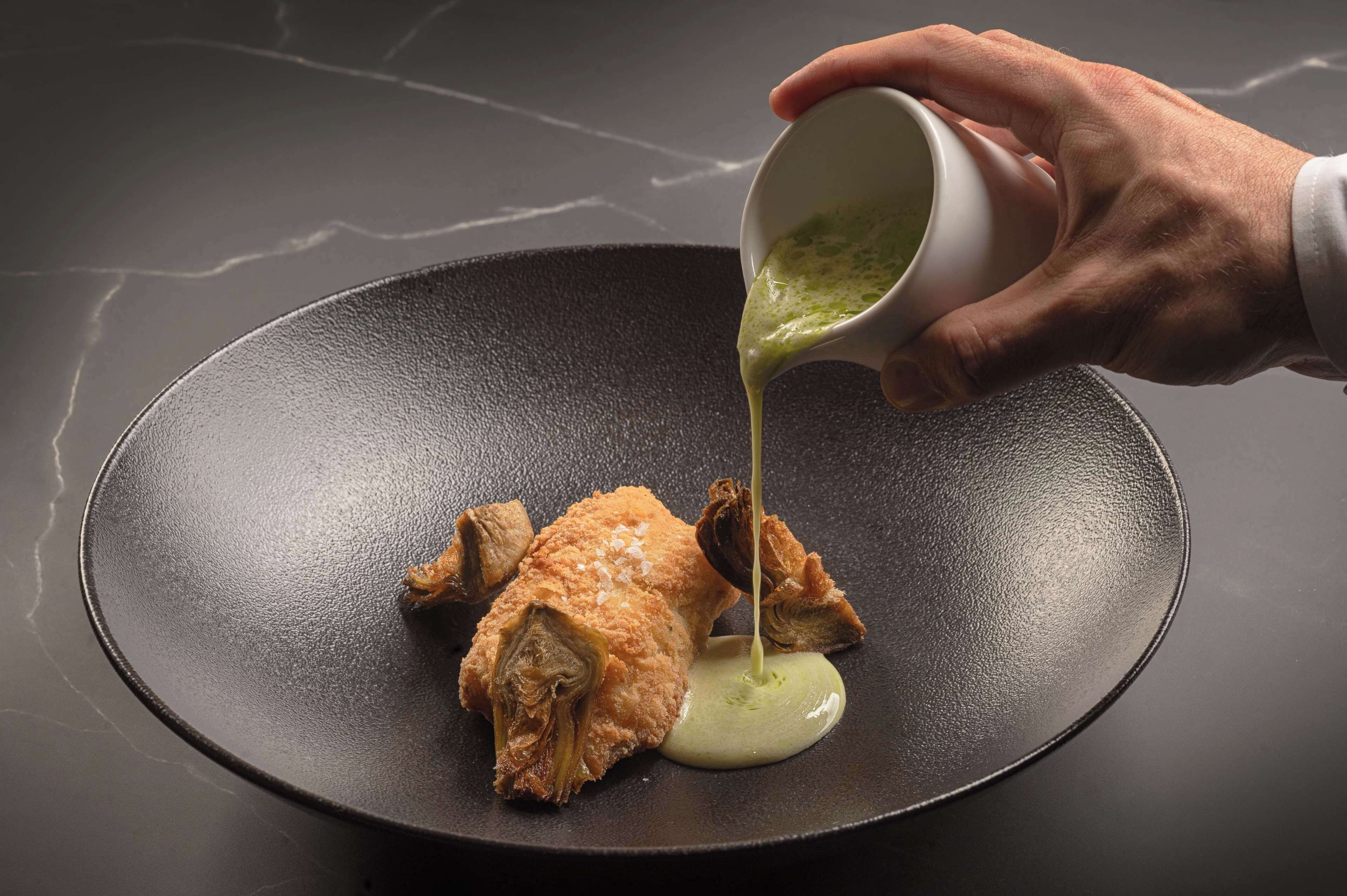 Una mano sirviendo salsa verde sobre comida frita en un plato negro.
