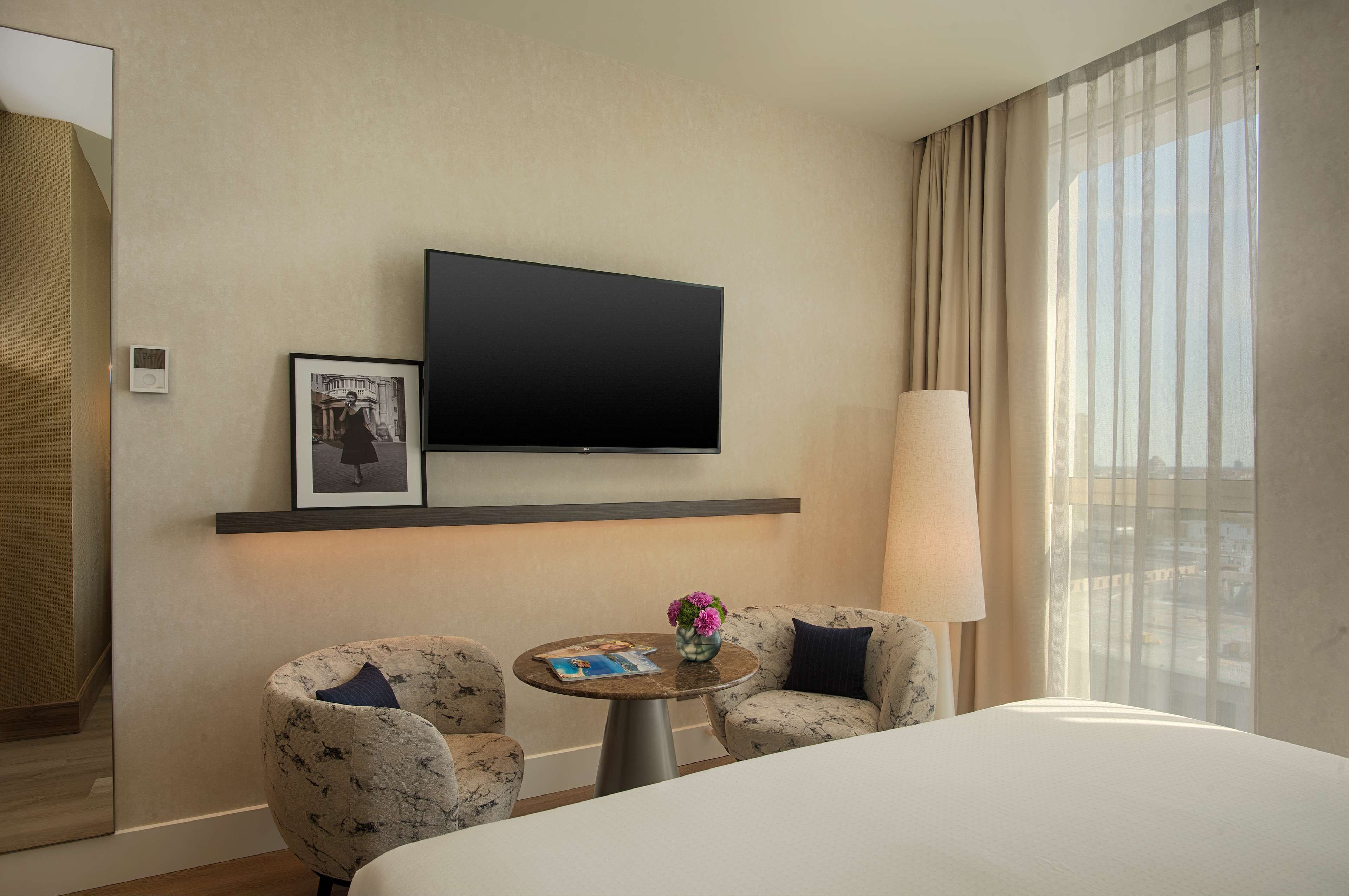 Habitación de hotel con cama, dos sillones, una mesita con un florero, televisor de pantalla plana montado en la pared y ventana con cortinas.