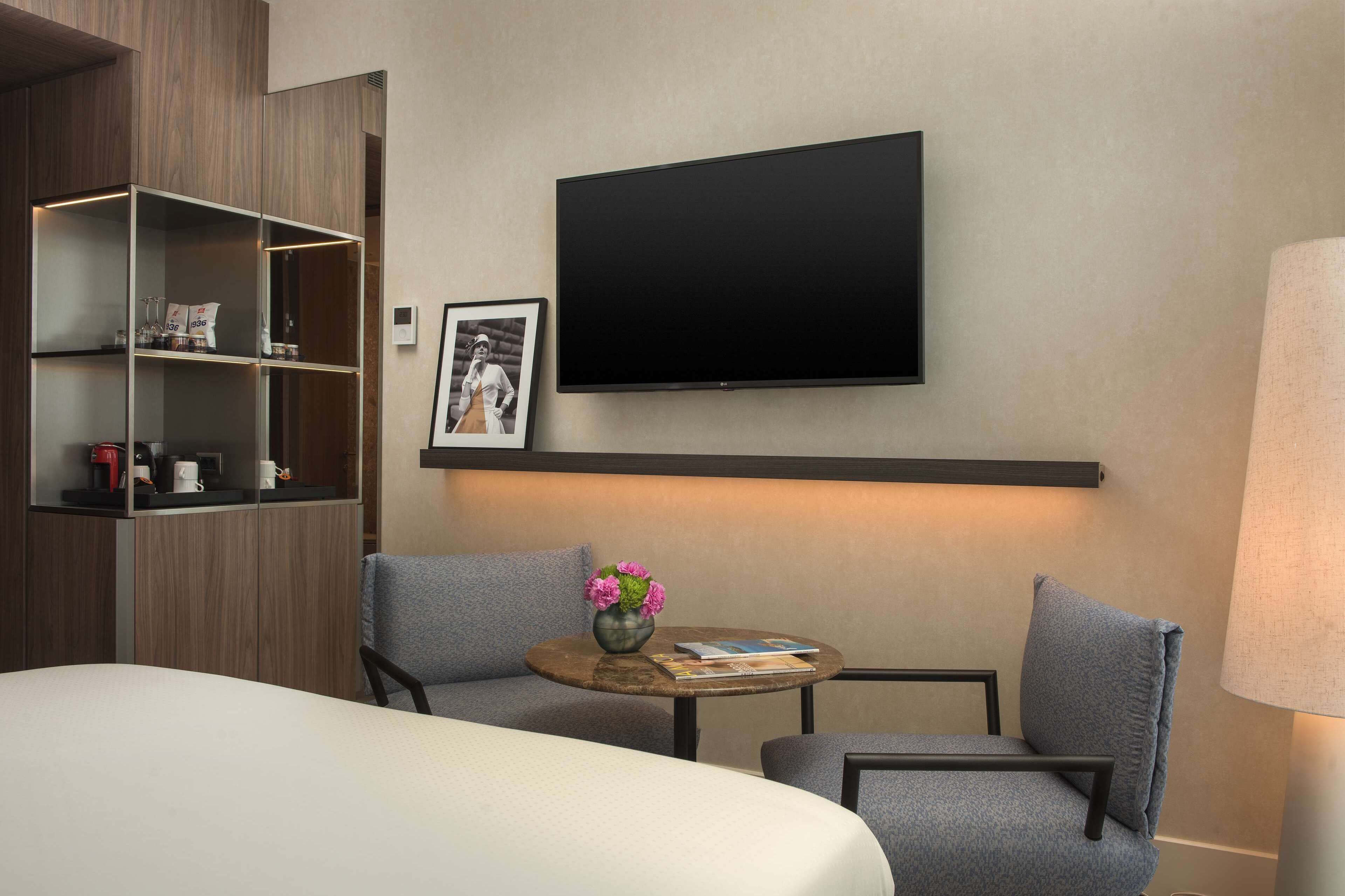 Ein Hotelzimmer mit einem Bett, zwei Sesseln, einem kleinen Tisch mit Blumenvase, einem flachbildfernseher an der Wand und einem Regal mit Gegenständen.