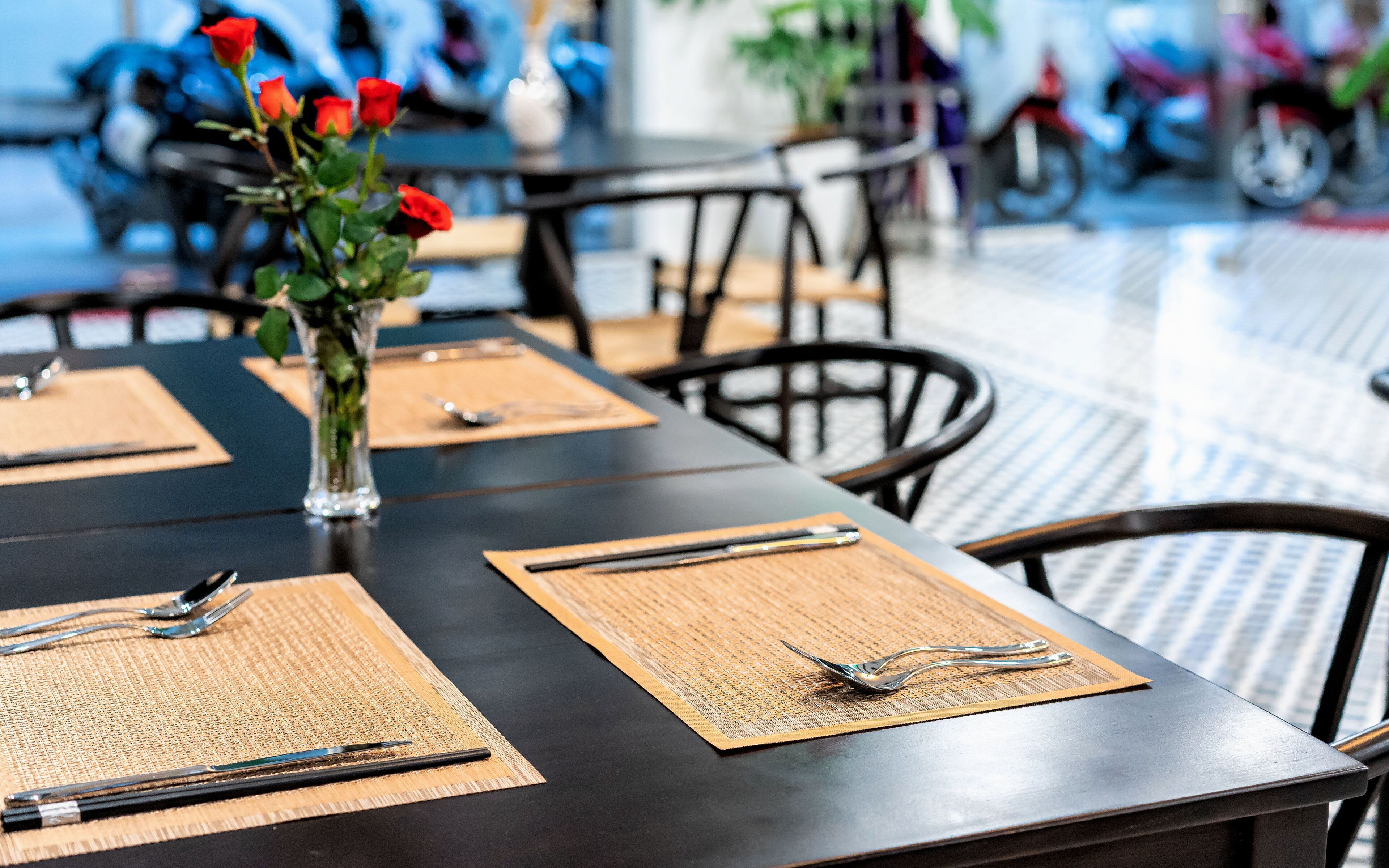 Una mesa de comedor dispuesta con manteles individuales, cubiertos y un florero con rosas rojas.