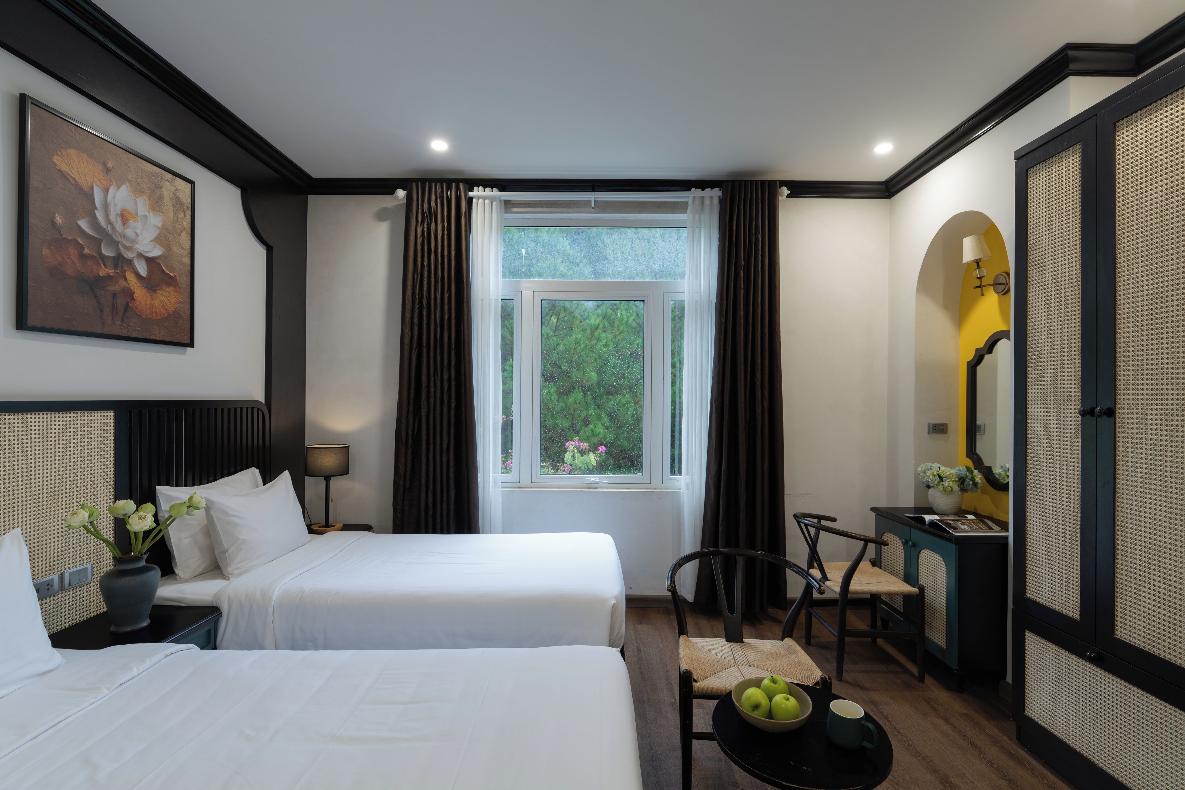 A hotel room with two beds, a desk, a chair, a window with curtains, a painting on the wall, and a lamp.