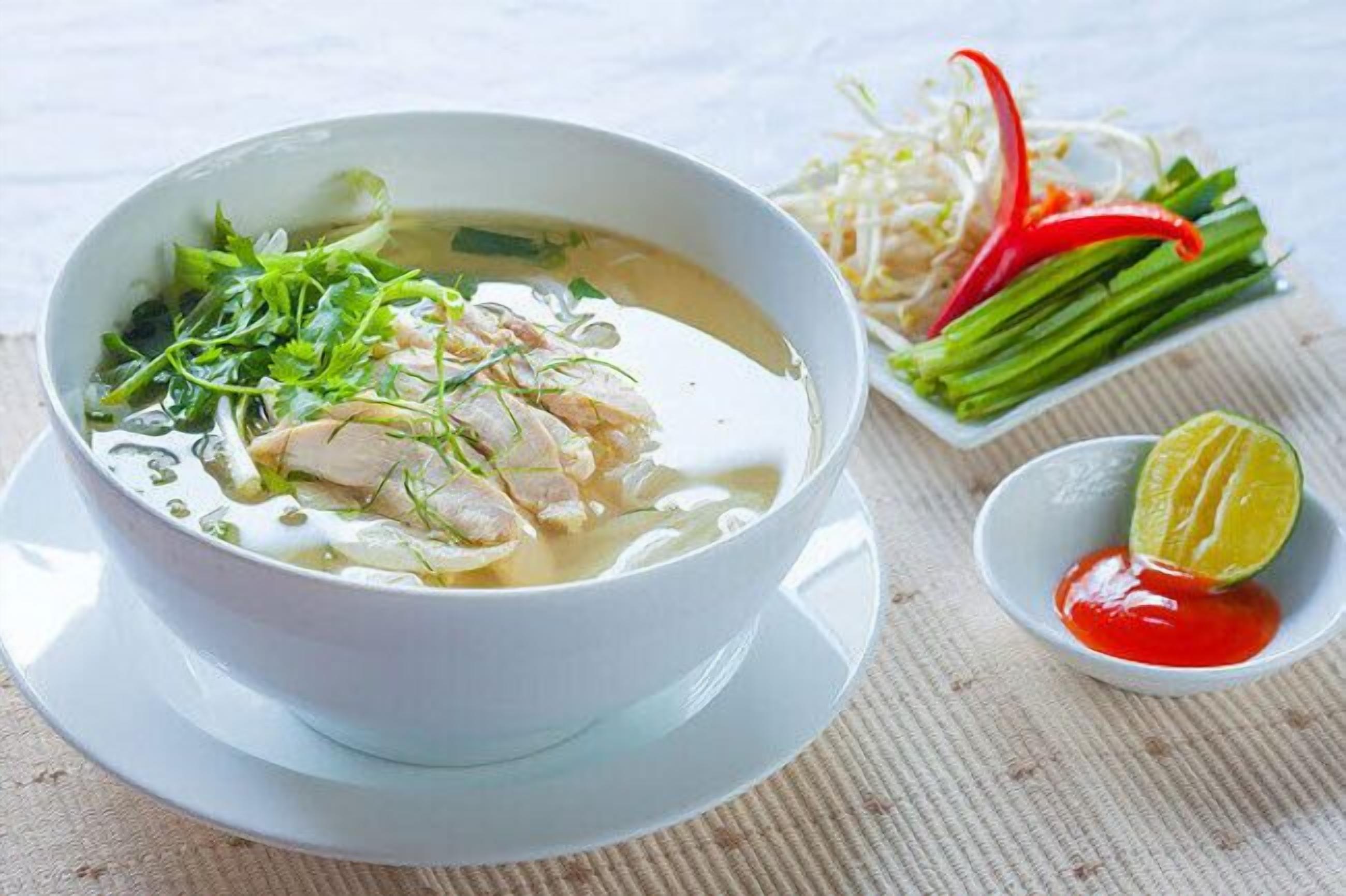 A bowl of soup with noodles and vegetables, accompanied by a small bowl of red sauce and a lime wedge.