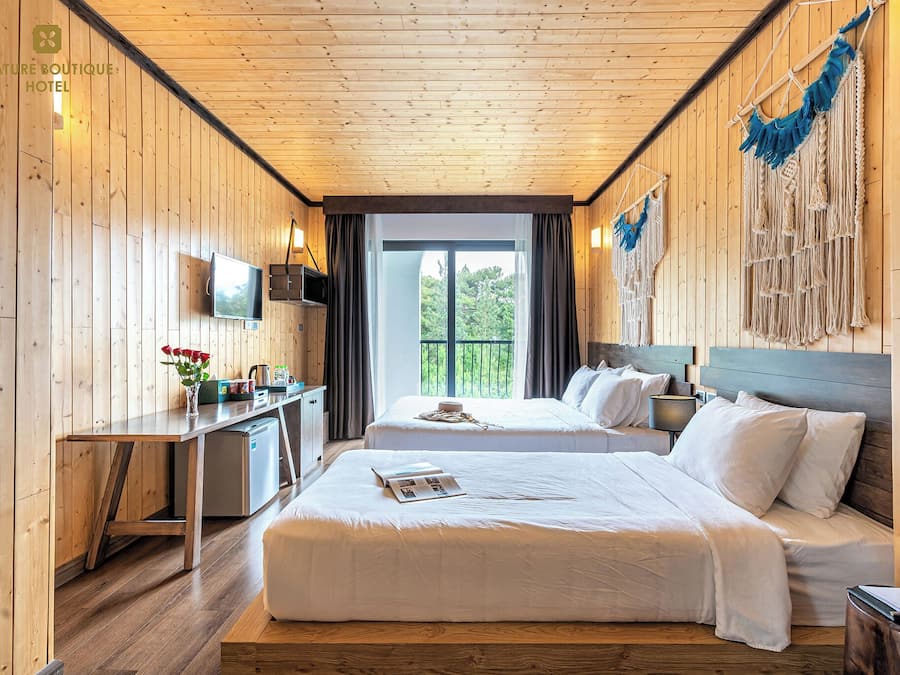 A hotel room with wooden walls and ceiling, two beds, a desk with a TV, and a balcony with a view of greenery.