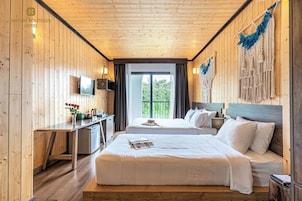 Quarto de hotel com paredes e teto de madeira, duas camas, uma escrivaninha com televisão e uma varanda com vista para vegetação.