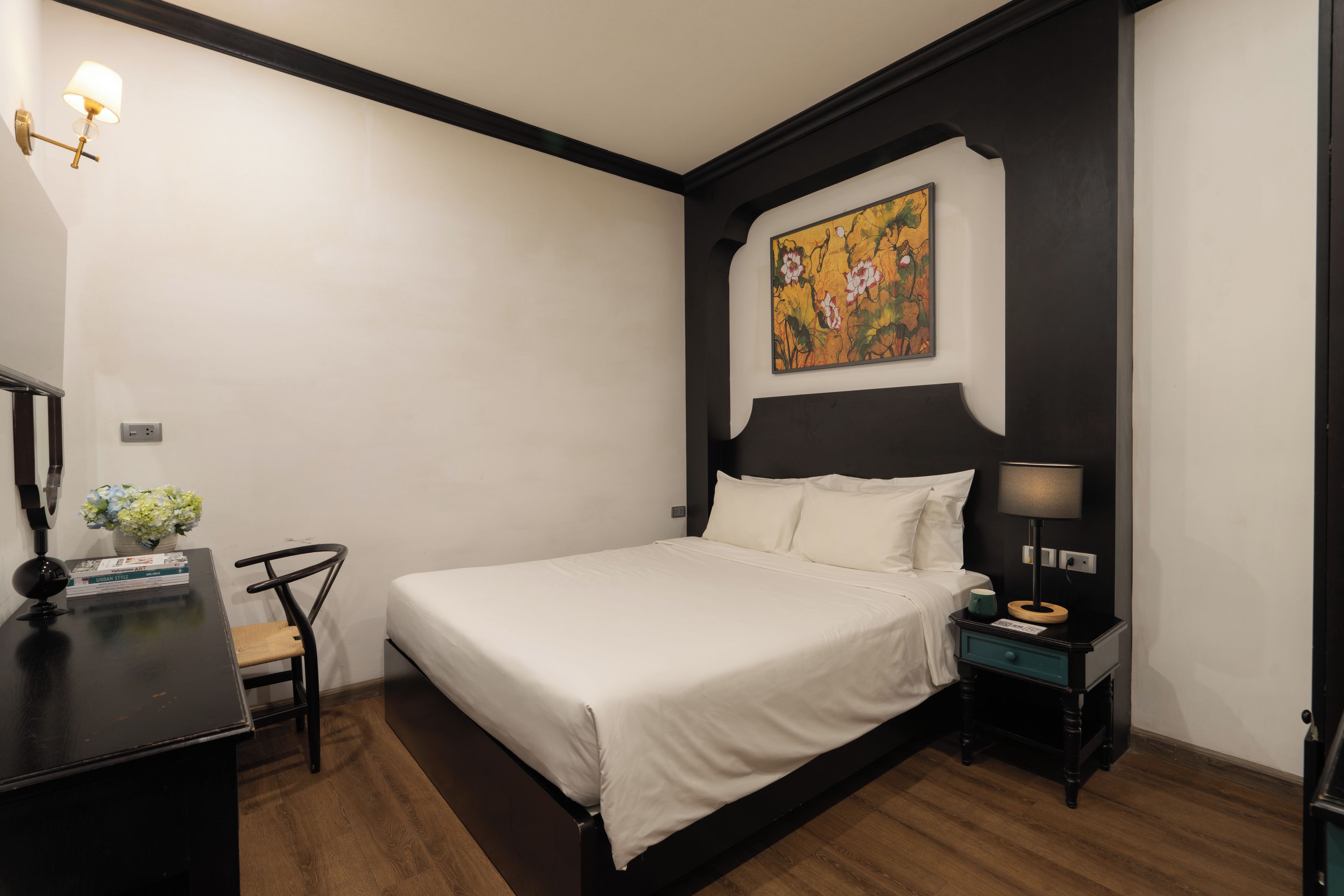 A hotel room with a bed, bedside table, lamp, chair, and a painting on the wall.