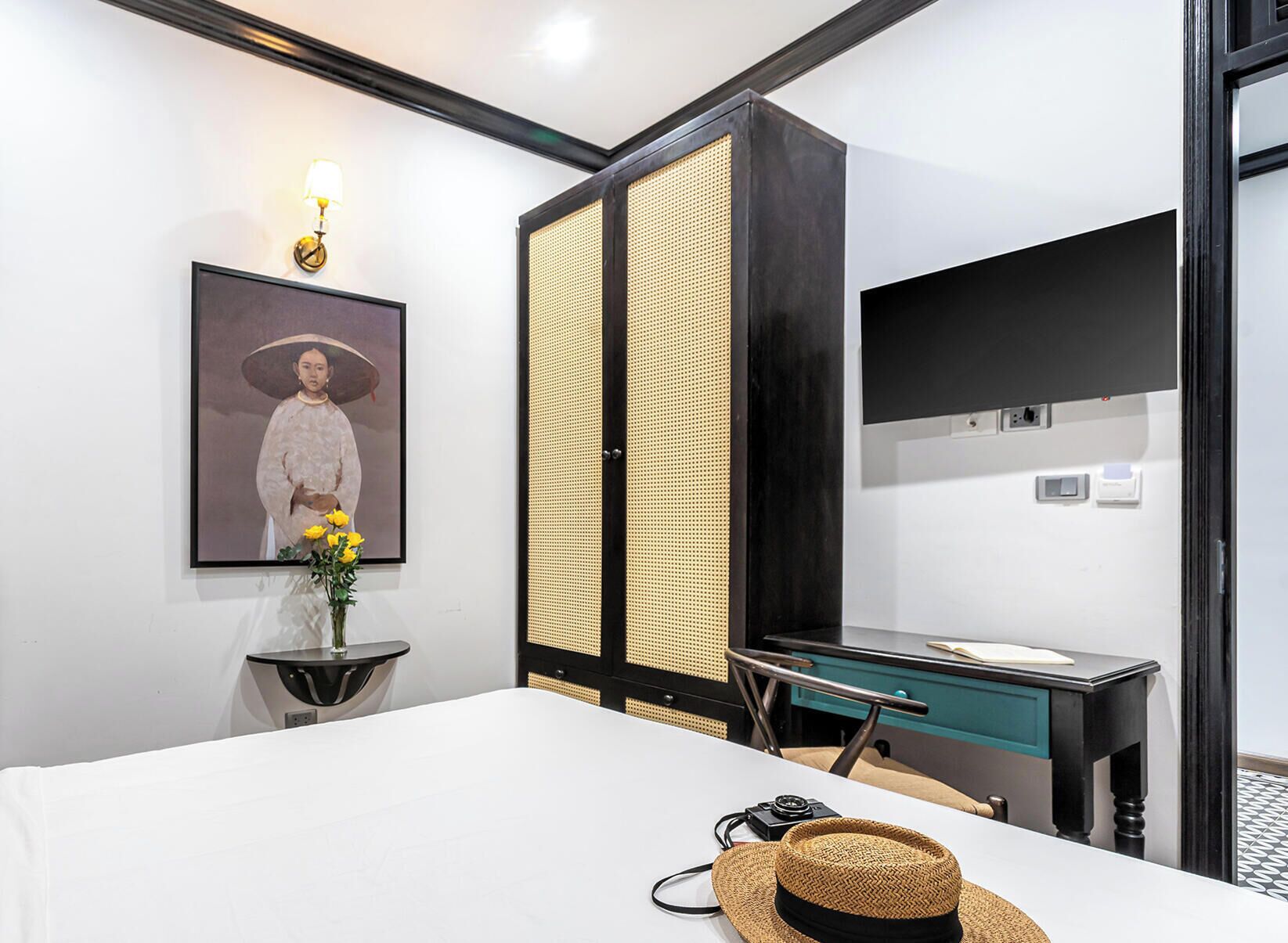 Una camera d'albergo con un letto, una scrivania, una sedia, uno specchio, un quadro incorniciato e un vaso con fiori.