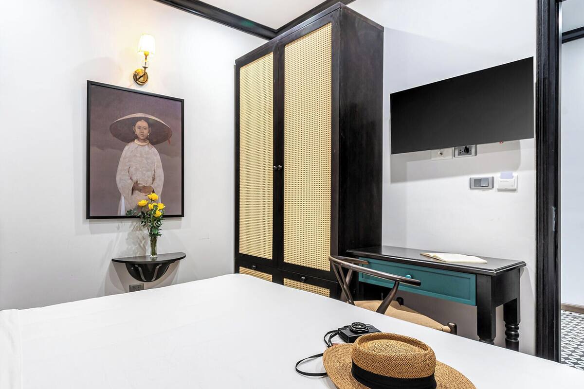 A hotel room with a bed, a desk, a chair, a mirror, a framed picture, and a vase with flowers.