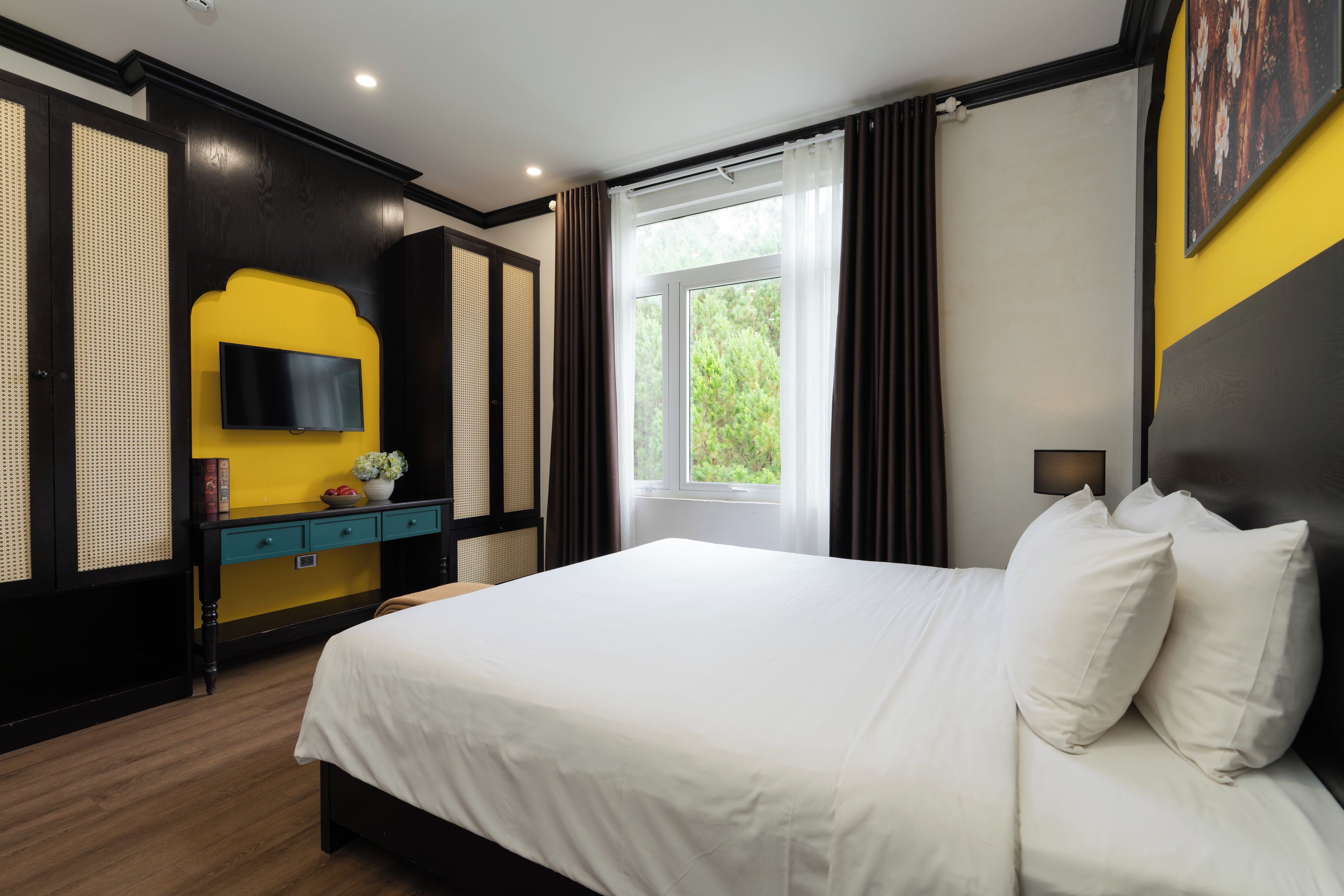 A hotel room with a bed, a TV mounted on a wall, a window with curtains, and a view of greenery outside.