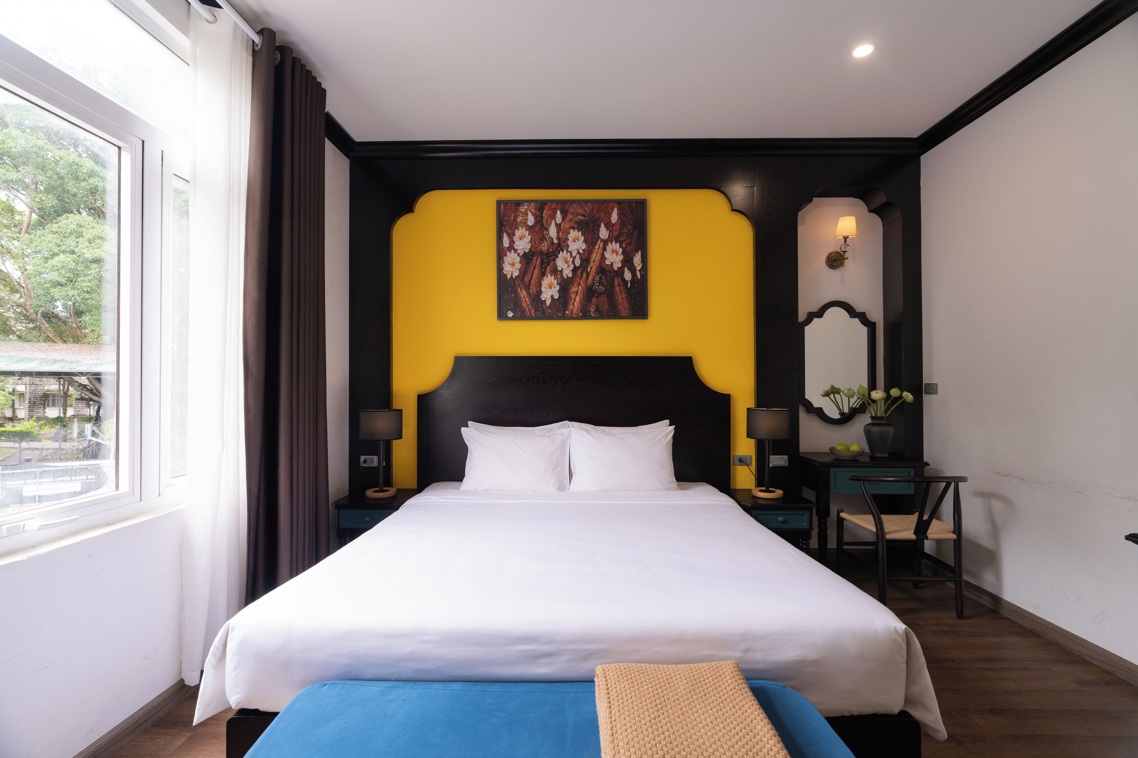 A hotel room with a large bed, a desk with a chair, a nightstand, a wall-mounted lamp, and a window with curtains.