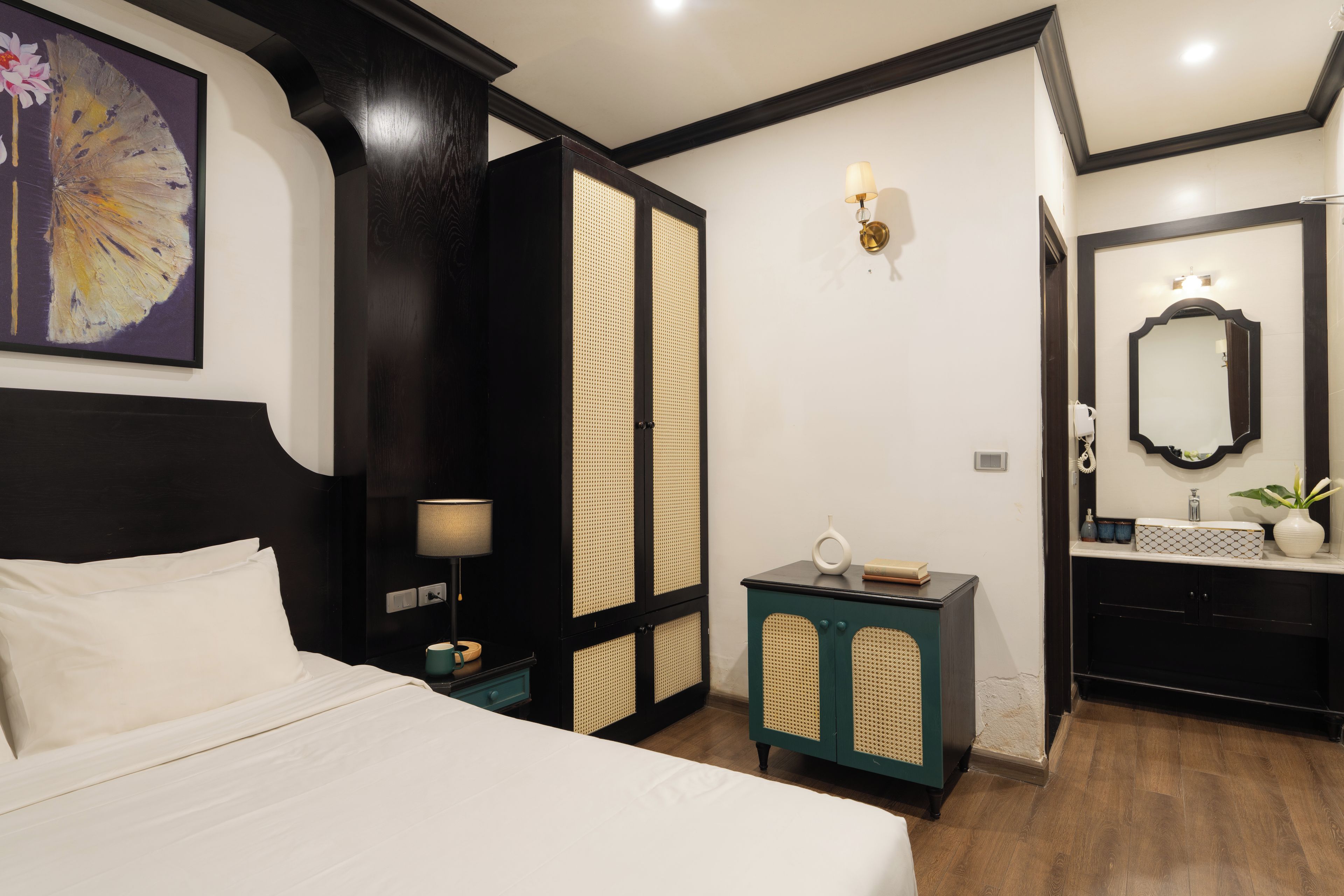 A hotel room with a bed, a nightstand, a teal cabinet, a bathroom with a mirror, and a wall-mounted light fixture.
