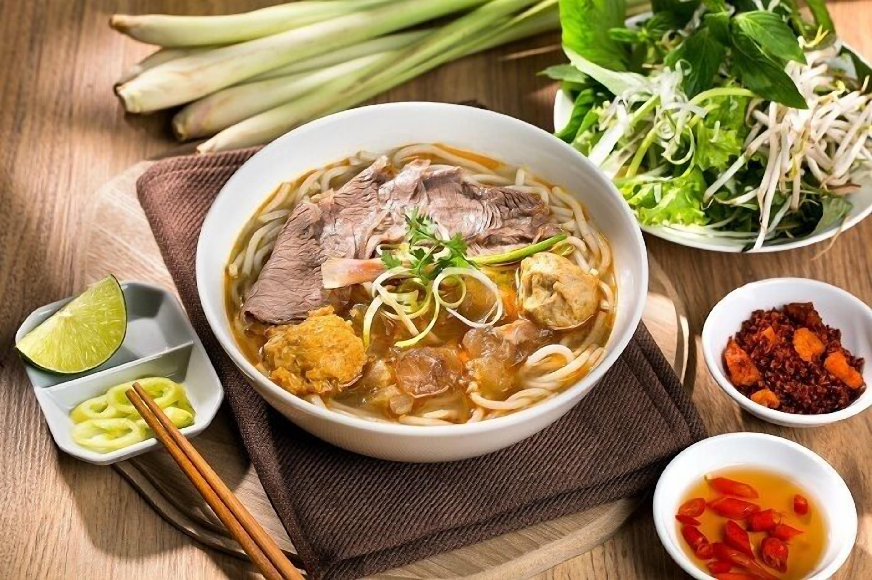A bowl of noodle soup with beef, tofu, and vegetables, accompanied by a side of fresh herbs, bean sprouts, and two small bowls of condiments.