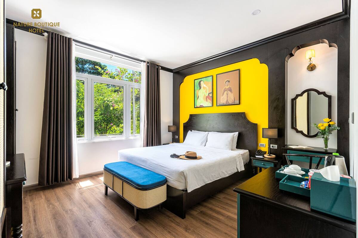 A hotel room with a large bed, a bedside table, a nightstand, a window with curtains, and a view of greenery outside.