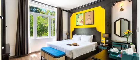 A hotel room with a large bed, a bedside table, a nightstand, a window with curtains, and a view of greenery outside.