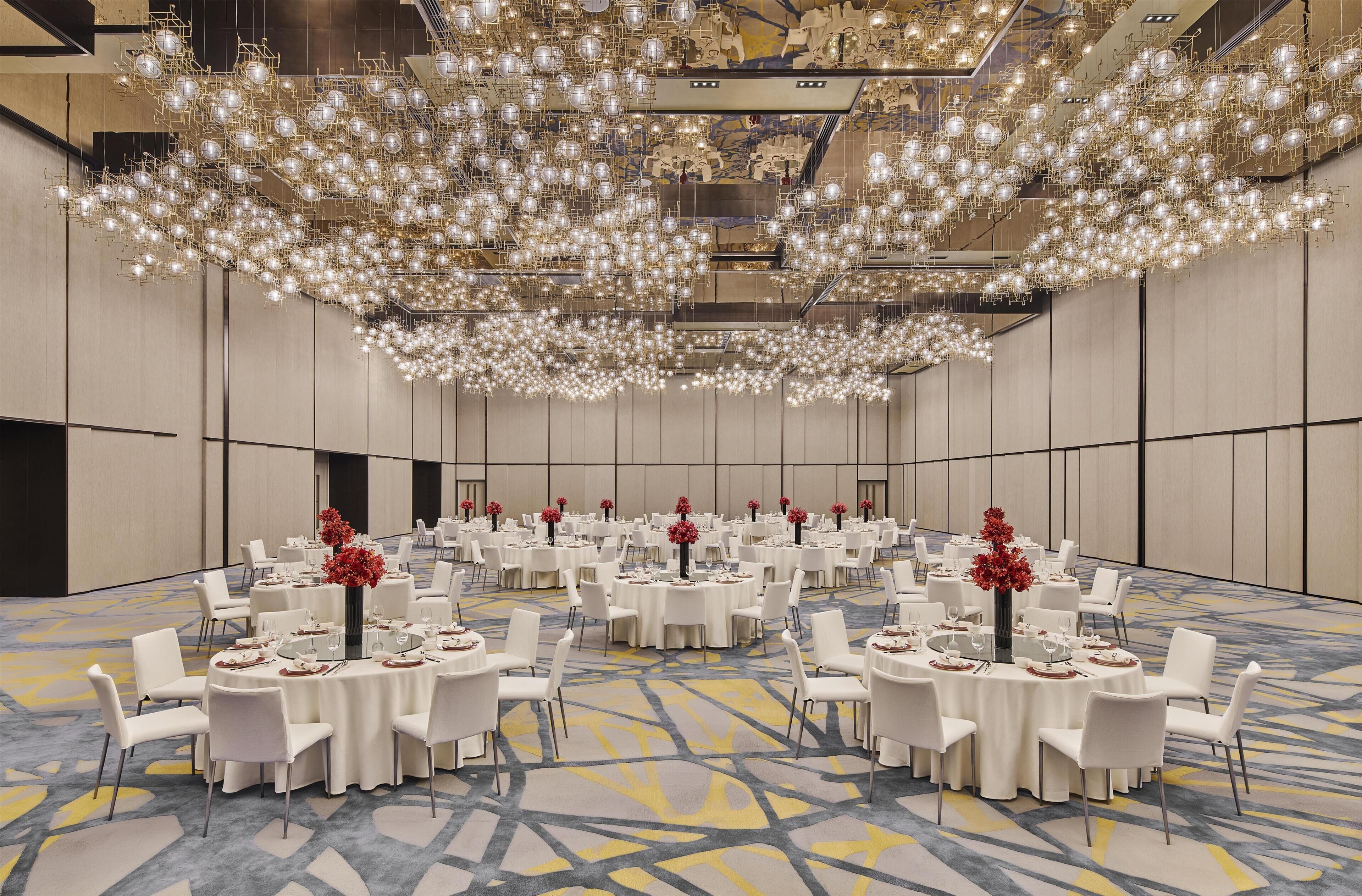Une grande salle de conférence dotée de tables rondes, de chaises blanches et d’un plafond orné de luminaires sphériques.