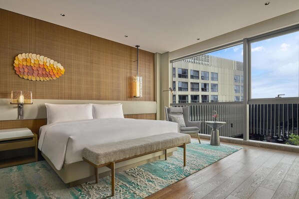Una habitación de hotel moderna con una cama grande, un pequeño espacio para sentarse, un balcón con vistas a la ciudad y una obra de arte decorativa en la pared.