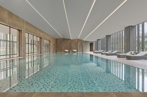 Una piscina interior moderna con paredes de vidrio transparente, paneles de madera y sillones de descanso a lo largo de los bordes.