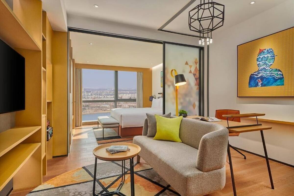 Une chambre d’hôtel moderne équipée d’un canapé, d’une petite table, d’un bureau, d’une télévision et d’une grande fenêtre donnant sur la ville.