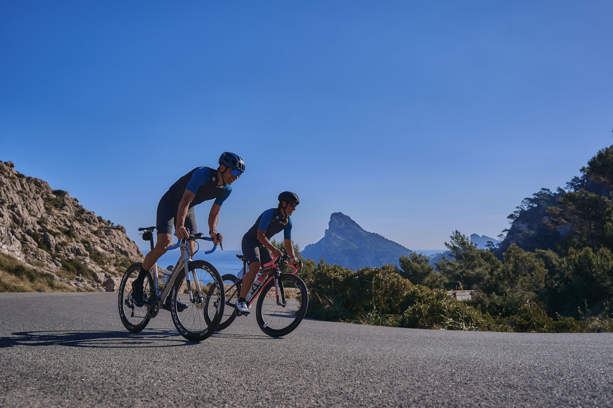 Dos ciclistas en una carretera pavimentada con montañas al fondo.