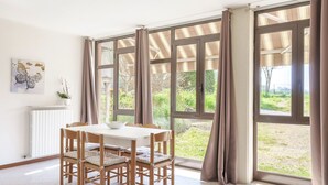 Een eetgedeelte met een tafel en stoelen, grote ramen voorzien van gordijnen, een radiator en een aan de wand bevestigde plant.